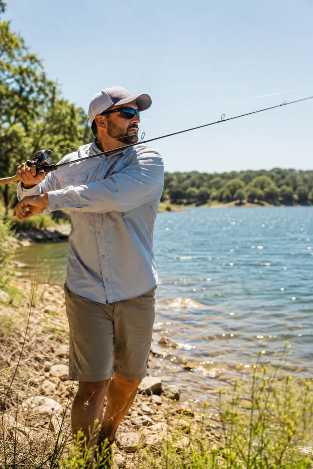 A man in a cap and polarized sunglasses casting a fishing rod from a lake shore