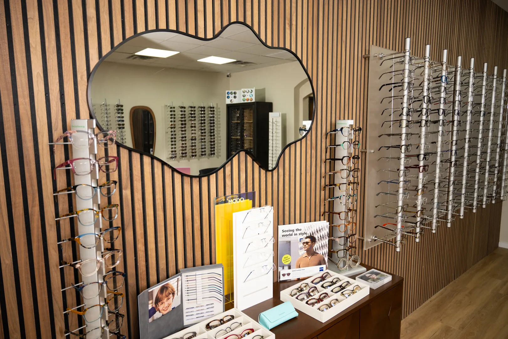 A wood-slat showroom wall of eyeglass frames beside a cloud-shaped mirror