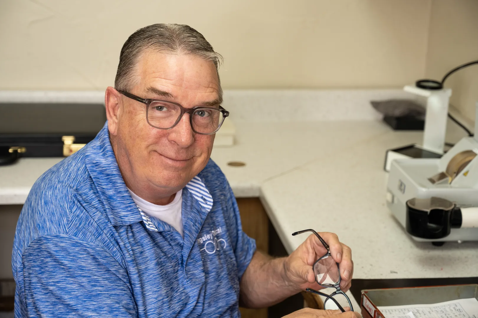 Robert Spurrier at the lab bench holding a pair of eyeglasses beside the edging equipment